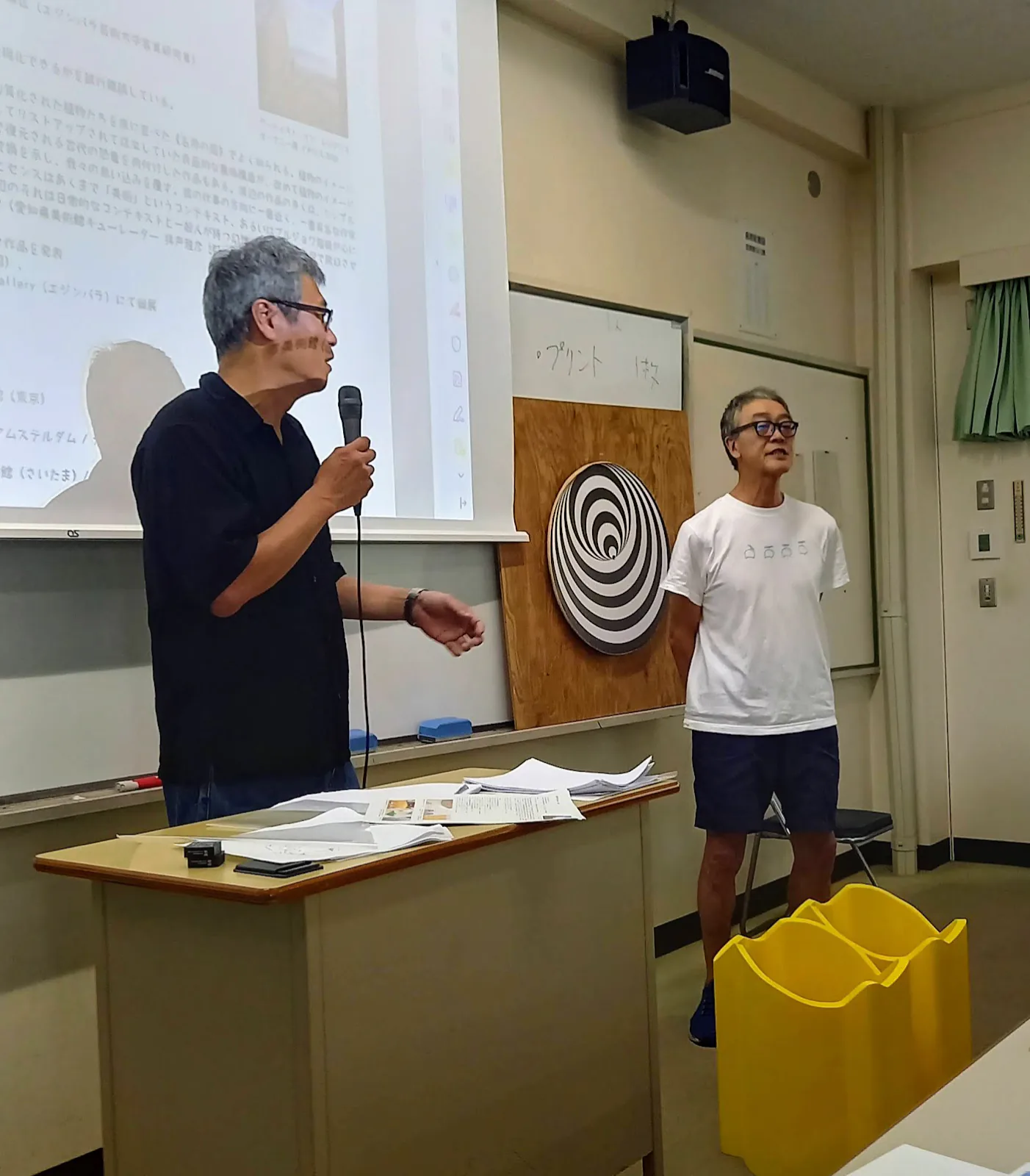愛知教育大学の教室でマイクを持つ渡辺英司と、錯視の円盤の横に立つ渡辺英治